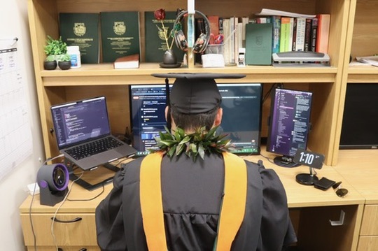 Taka Khoo at his lab desk in graduation robes, with his master’s hood and a lei